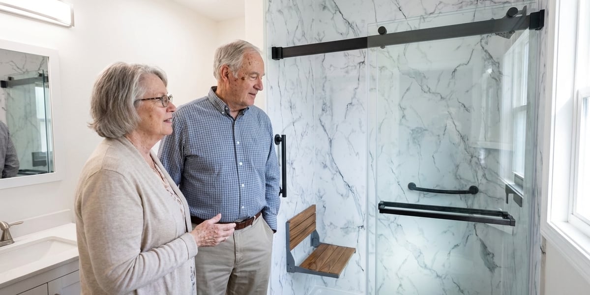 An older couple looking into their remodeled bathroom, at a walk-in shower with a grab bar and fold-down seat